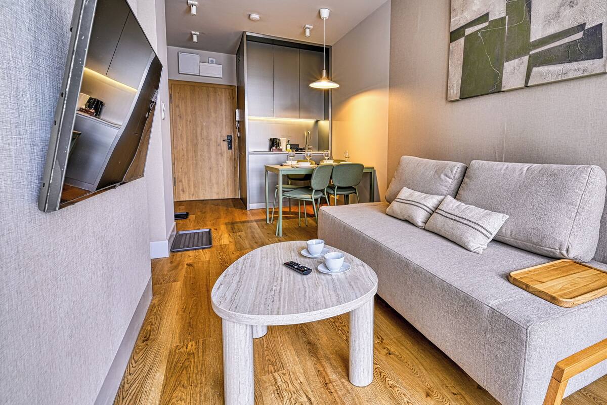 Ein modernes Wohnzimmer mit einem grauen Sofa, einem runden Couchtisch und einem Holznachttisch. Im Hintergrund ist eine Essecke mit einem grünen Tisch und Stühlen zu sehen.