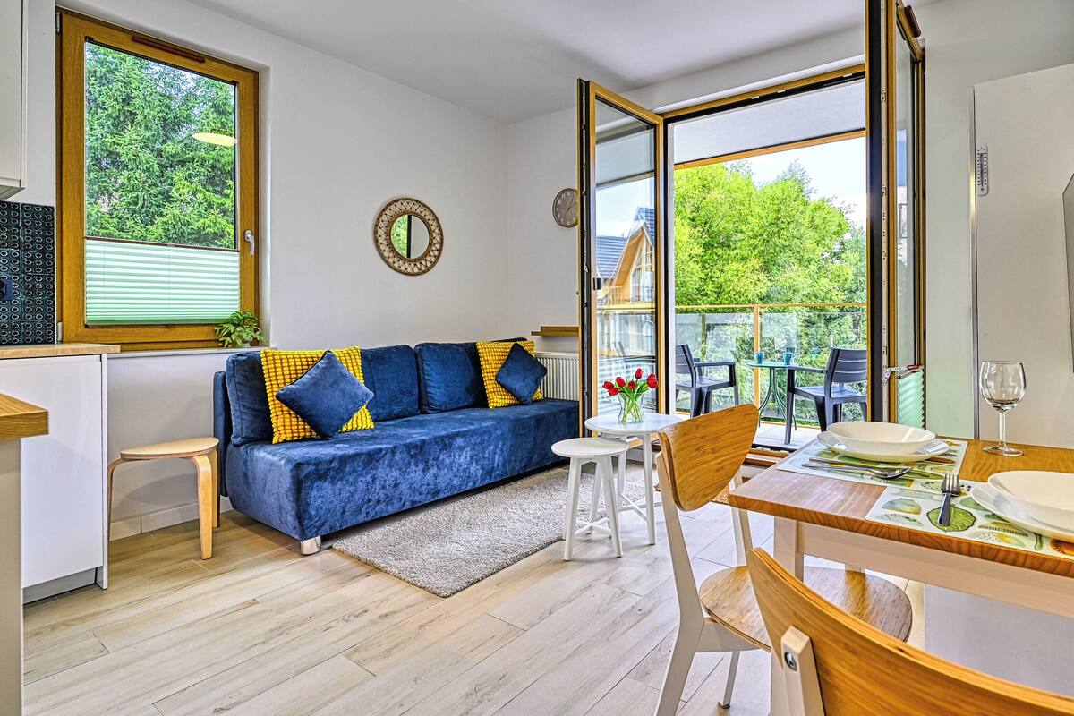 Ein modernes Wohnzimmer mit einem blauen Sofa, einem Esstisch mit Stühlen und einem großen Fenster, das einen Blick ins Grüne bietet.
