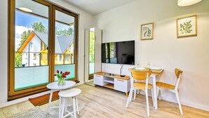 Un salon moderne avec un téléviseur, une table à manger et des chaises, de grandes fenêtres donnant sur des maisons et des œuvres d’art accrochées au mur.