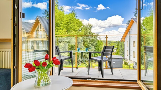 Un balcon avec une table et des chaises, un vase rempli de fleurs rouges et une vue sur des maisons et des arbres.