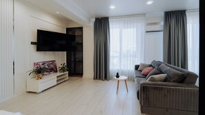 Un salon moderne avec un téléviseur à écran plat, des rideaux gris, un canapé gris et une petite table d’appoint blanche.