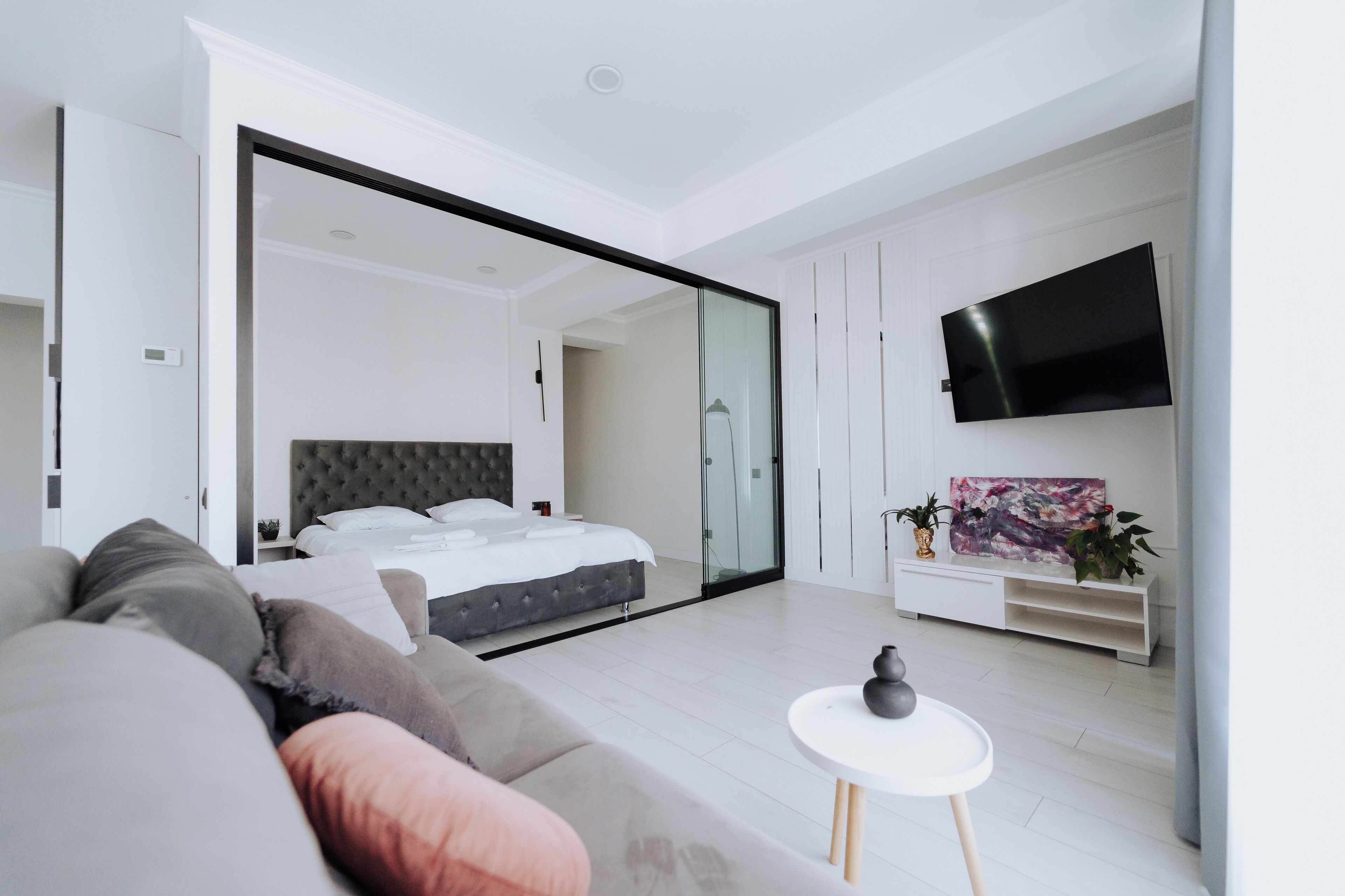 A modern bedroom with a large bed, sliding glass door to a bathroom, a flat-screen TV, and a white console table with plants.