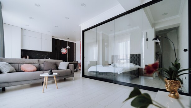 Un salon moderne avec un canapé gris, une petite table blanche et une cloison en verre donnant sur une chambre à coucher.