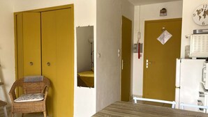 Ein Zimmer mit einem Holztisch, einem Korbstuhl, einem gelben Wandschrank, einer weißen Tür und einem weißen Kühlschrank.