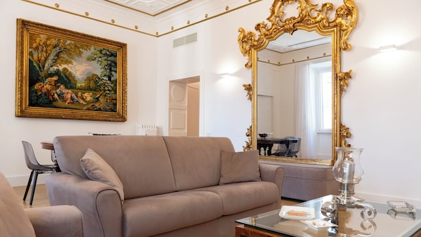 Un salon moderne avec un canapé beige, une table basse en verre, un grand miroir décoratif et un tableau représentant un paysage classique accroché au mur.