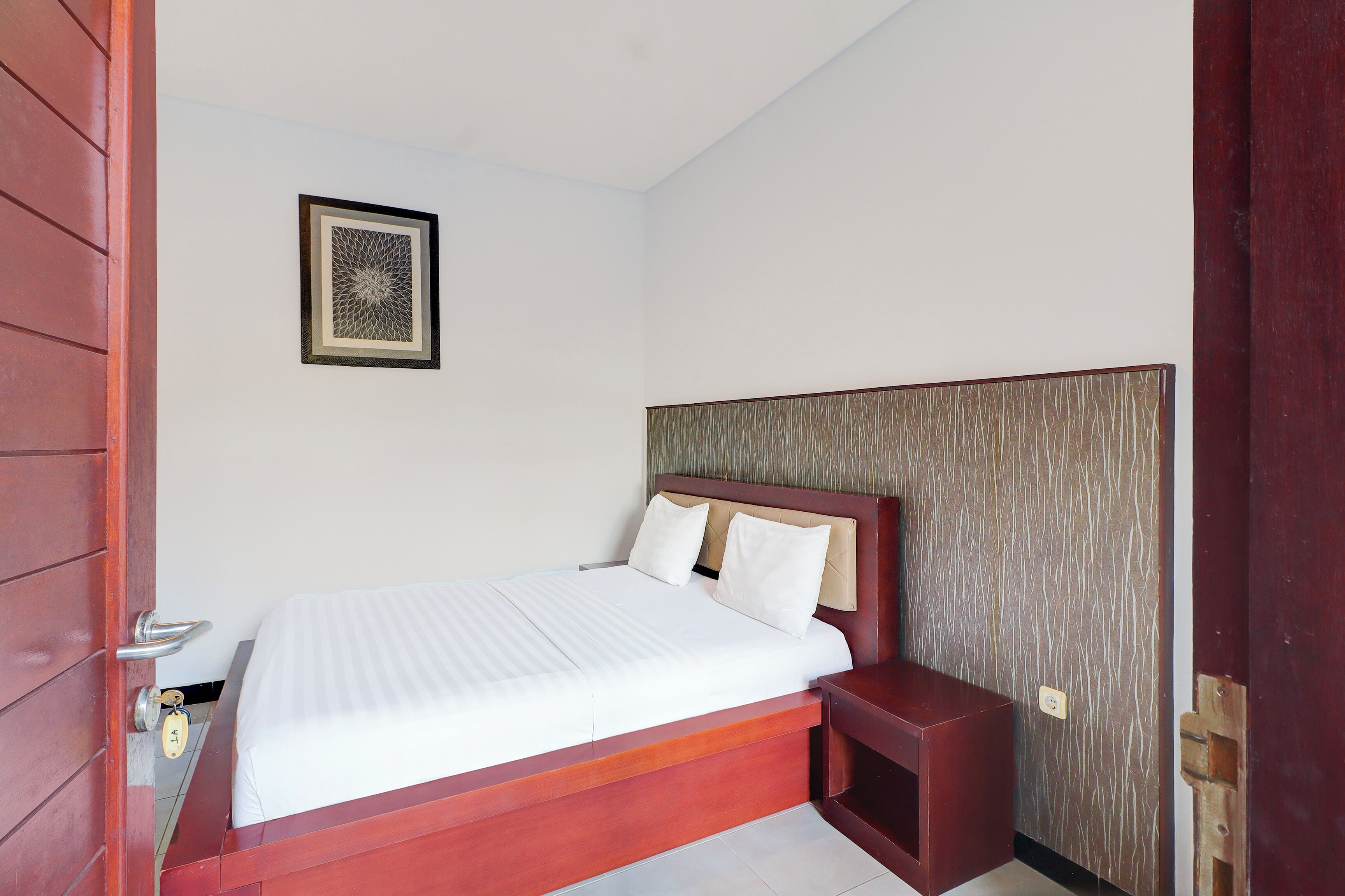 A neatly made bed in a hotel room with a wooden headboard, a small bedside table, and a framed picture on the wall.