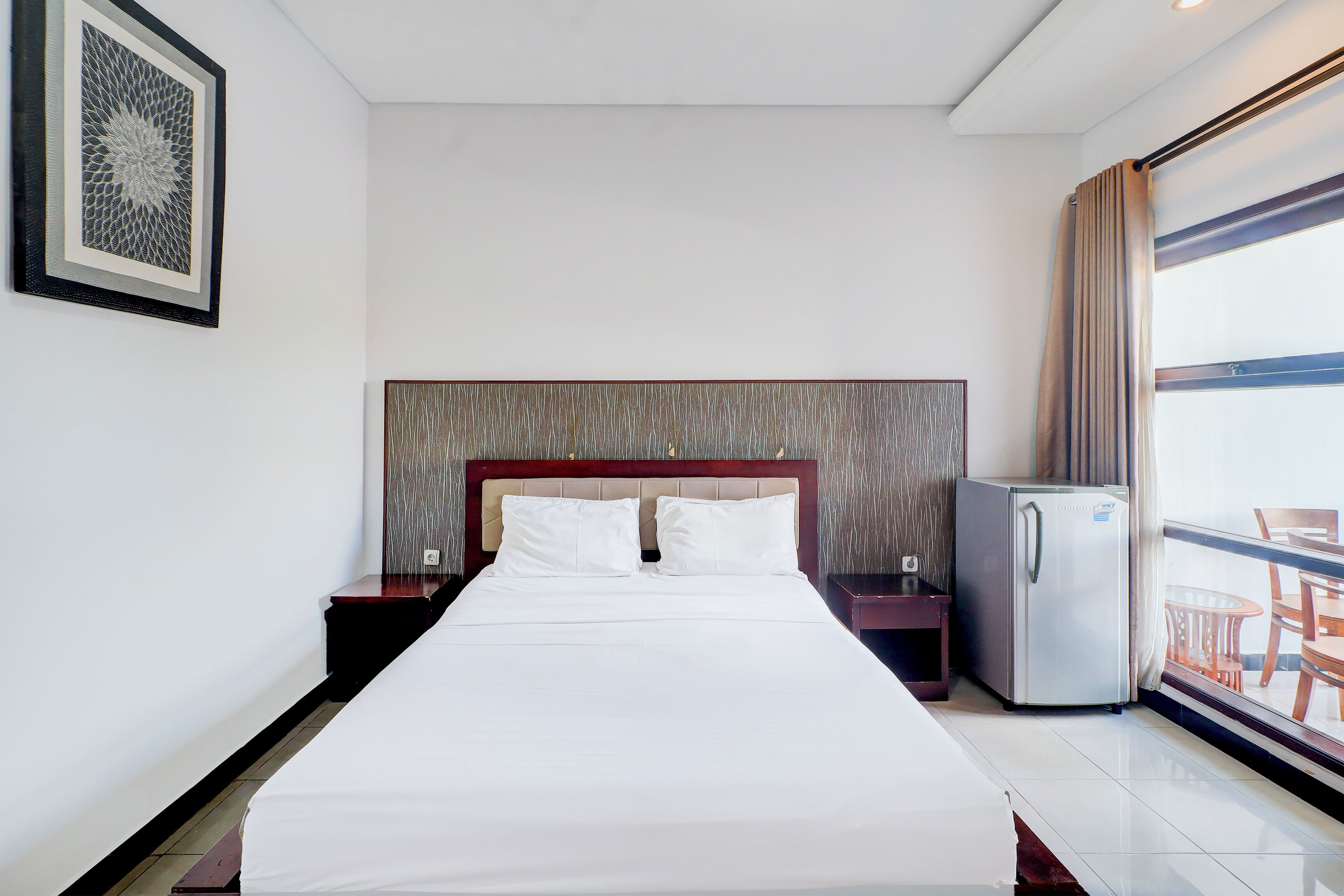 A hotel room with a large bed, a small refrigerator, a nightstand, and a window with a view of the outside.
