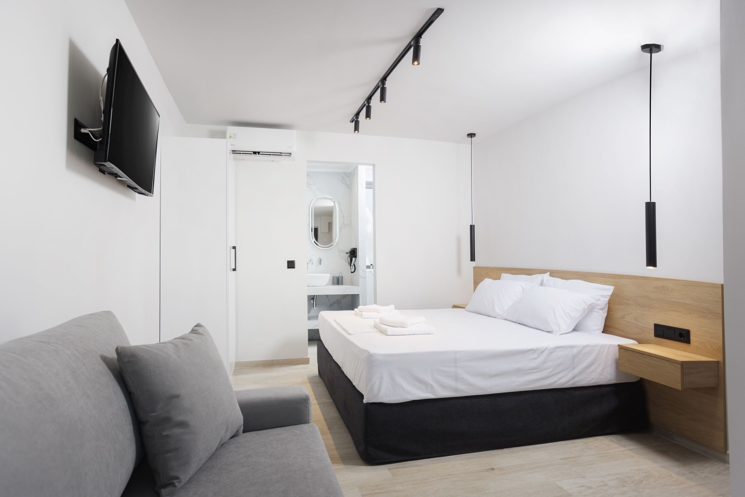 A modern hotel room with a bed, a grey sofa, a TV mounted on the wall, and a bedside table.