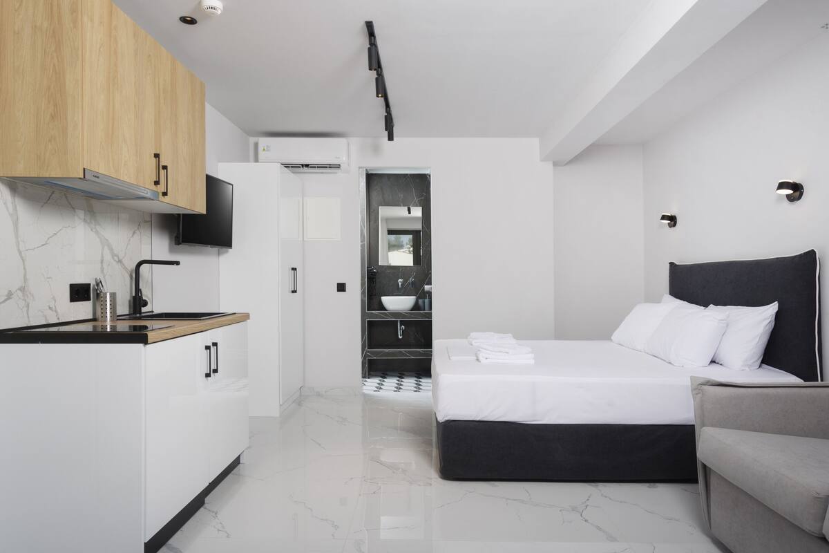 Una habitación de hotel moderna con zona de cocina, cama con ropa blanca y sofá gris.