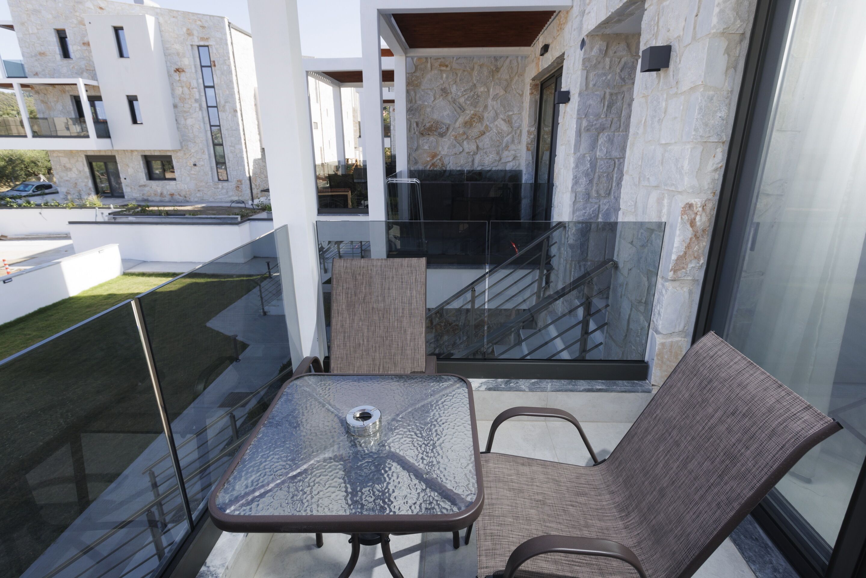 A balcony with a glass table and two chairs, a stone wall, and a view of neighboring buildings.
