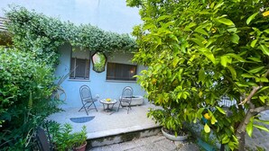 A courtyard with two chairs and a table, surrounded by lush greenery and a blue building. - Cleo Limone (Falconara Marittima)