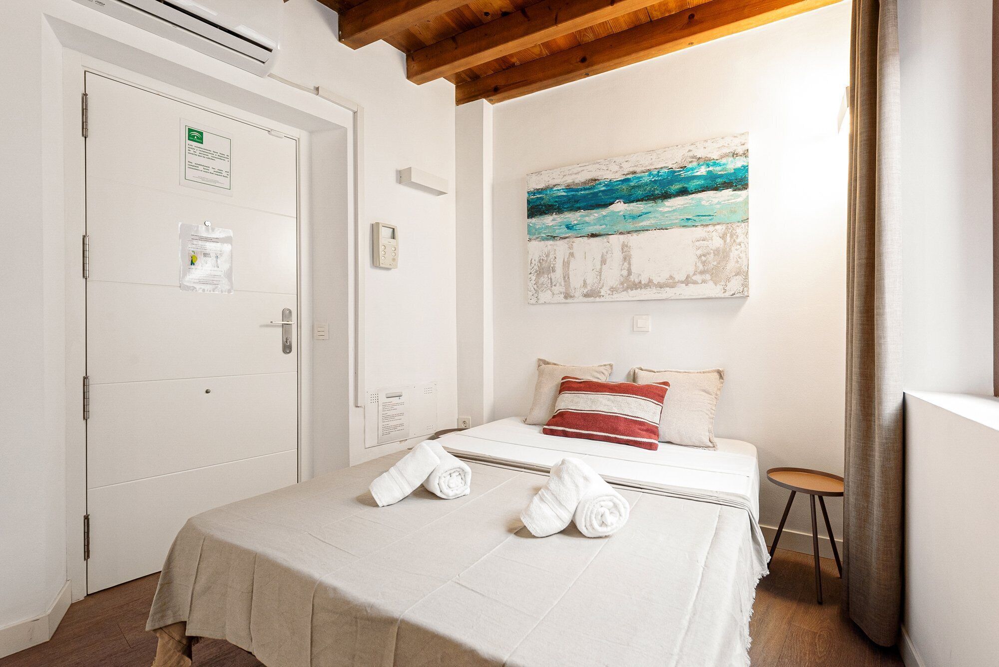 A hotel room with a bed, a painting, a door with a sign, and a small stool.
