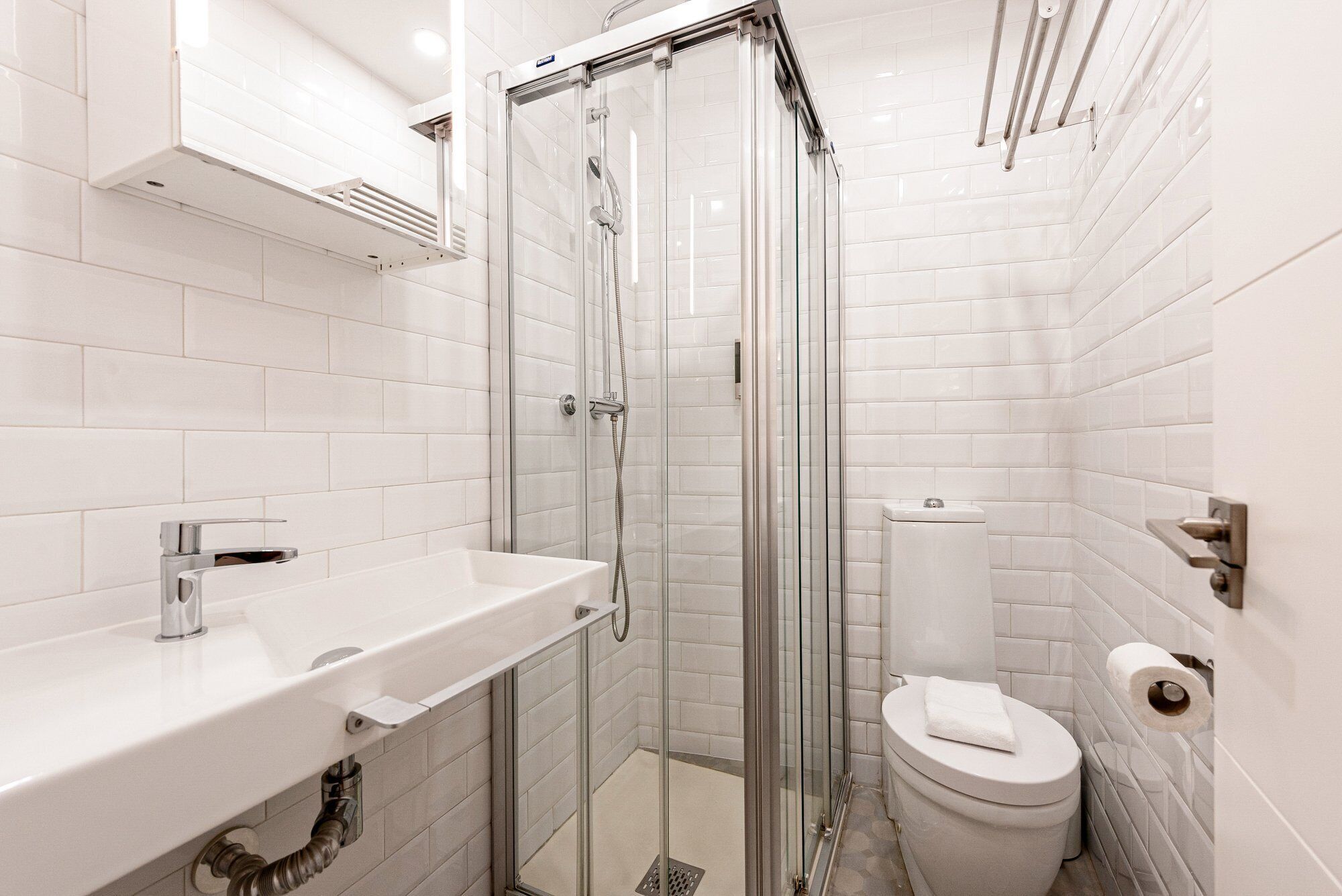 A modern bathroom with white tiles, a glass-enclosed shower, a white sink, and a toilet.