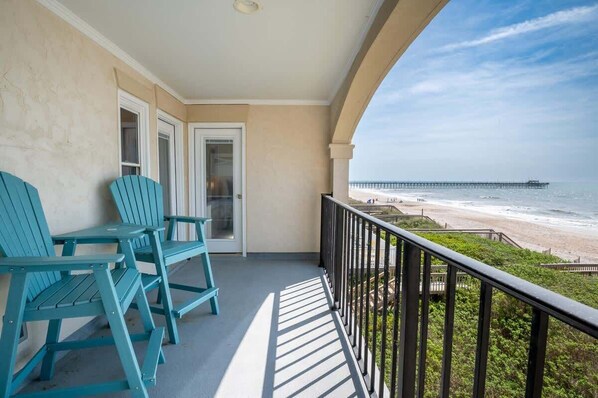 Un balcón con dos sillas Adirondack turquesa, vista a la playa y un muelle a lo lejos.