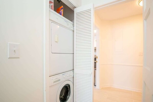 A compact laundry area with a front-loading washer and dryer, a small storage shelf above the dryer, and a door leading to another room.