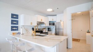 A modern kitchen with white cabinetry, a marble countertop, and stainless steel appliances. There are three white bar stools and a built-in island.