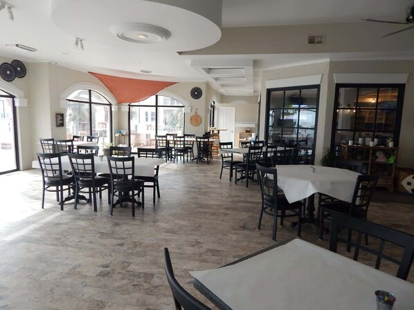 A spacious restaurant with multiple tables and chairs, large windows, and a modern ceiling design.