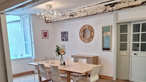 Comedor con mesa de madera, sillas blancas, una araña y pared rústica con espejo y obras de arte.