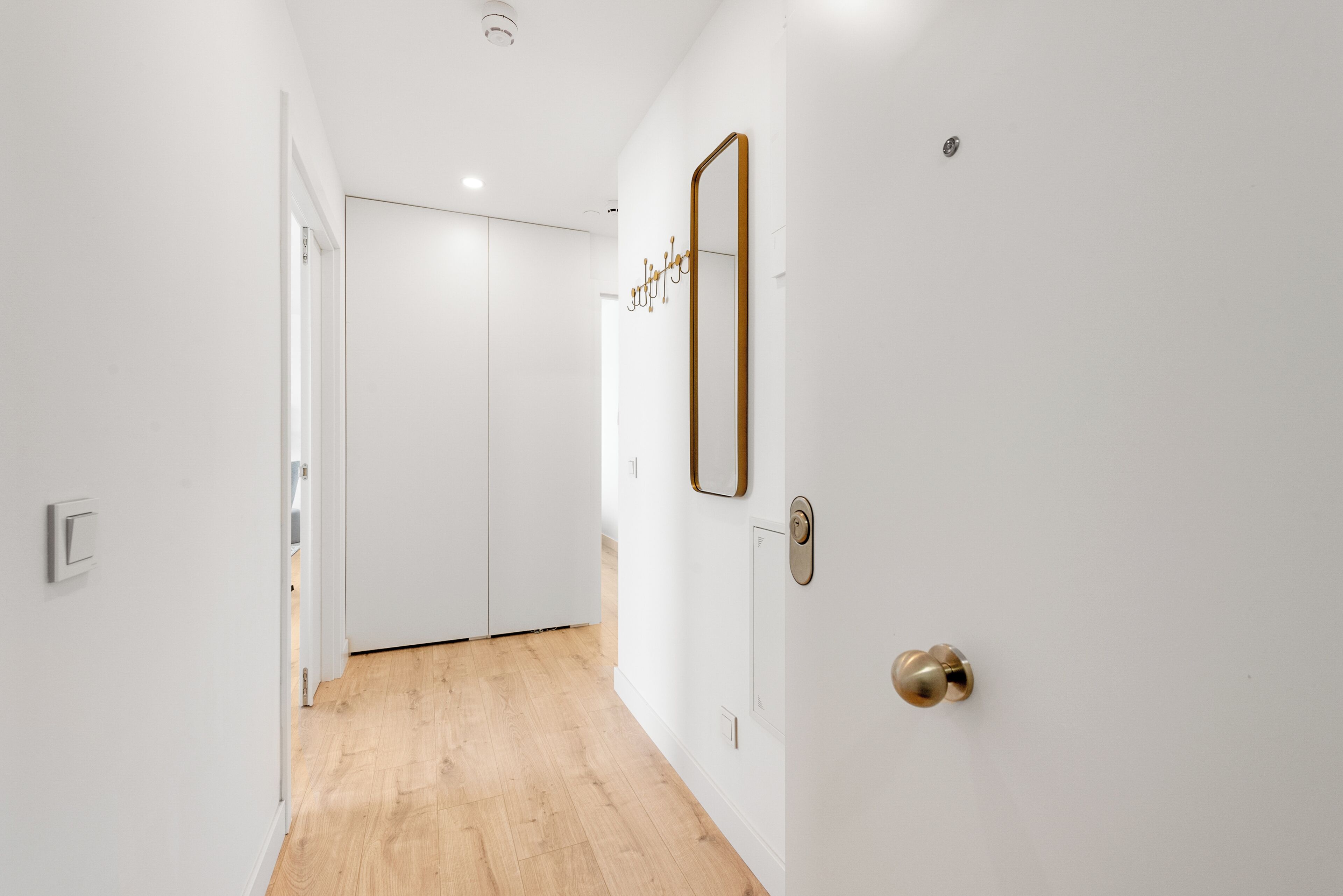 A hallway with a wooden floor, white walls, a door with a gold handle, and a mirror on the wall.
