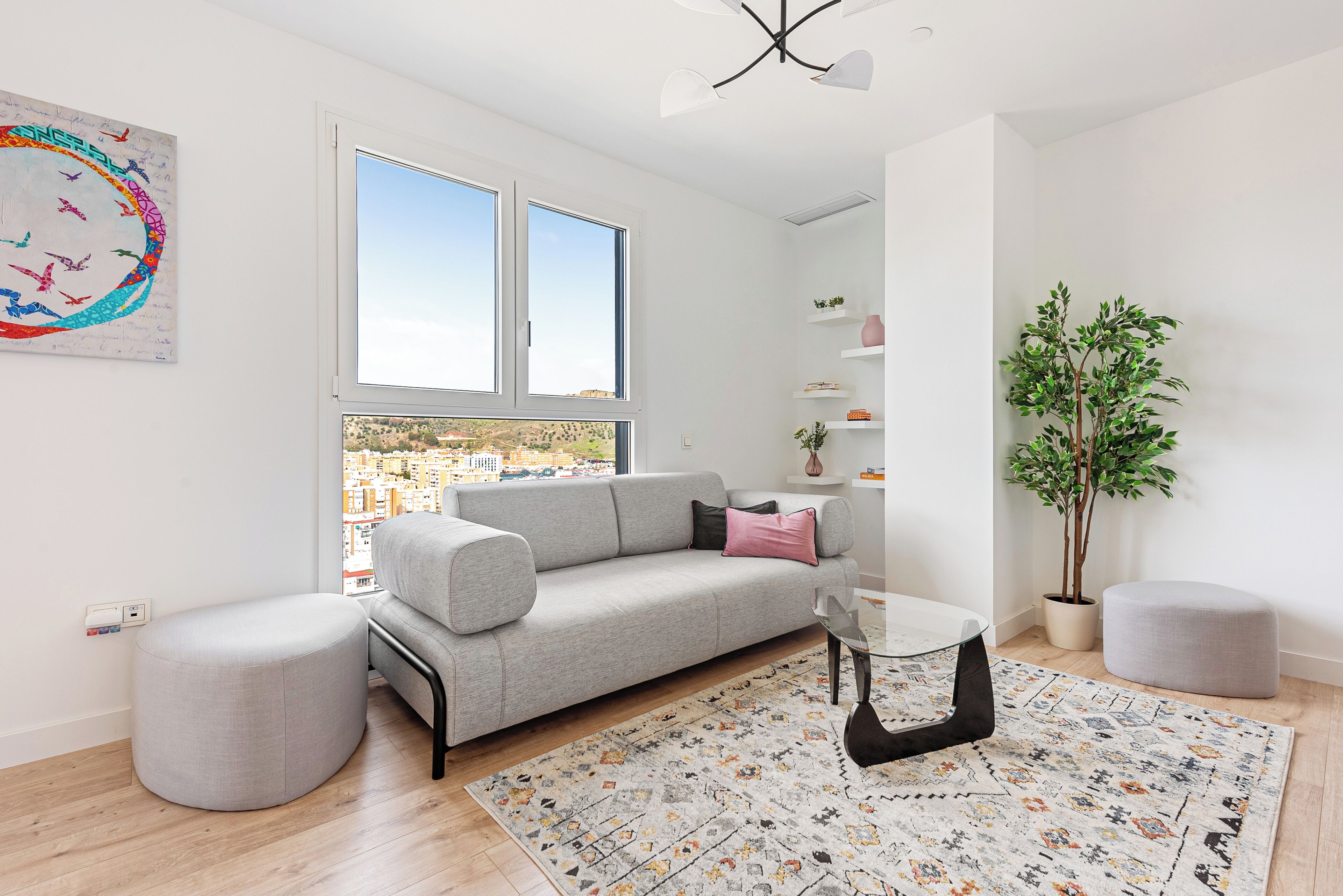 Una sala moderna con un sofá gris, un banquete redondo y una mesa de centro de vidrio. Hay un ventanal con vistas a la ciudad, una planta en maceta y un estante montado en la pared con objetos decorativos.