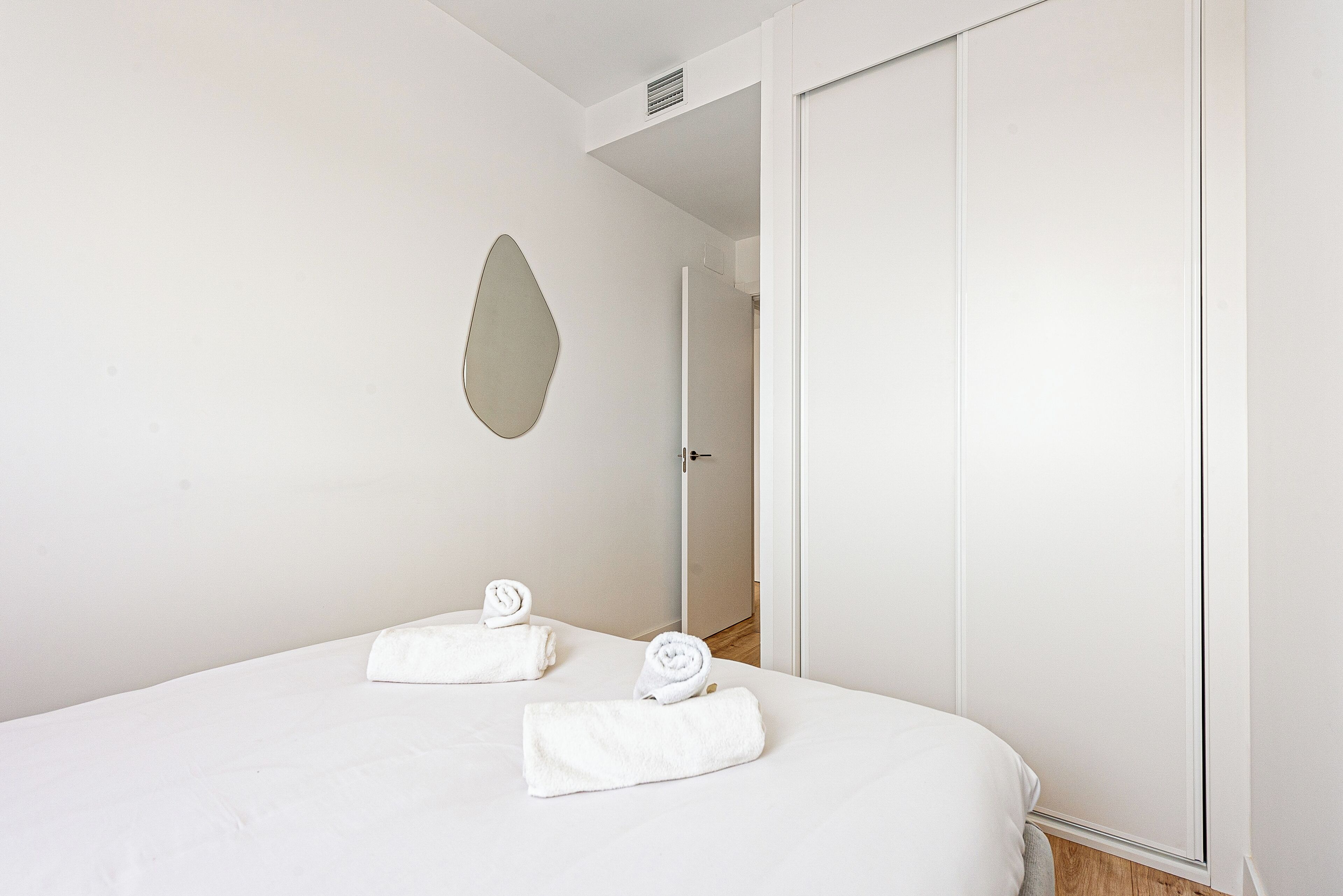 Quarto de hotel com uma cama, duas toalhas enroladas, um espelho e uma porta que dá para outro cômodo.