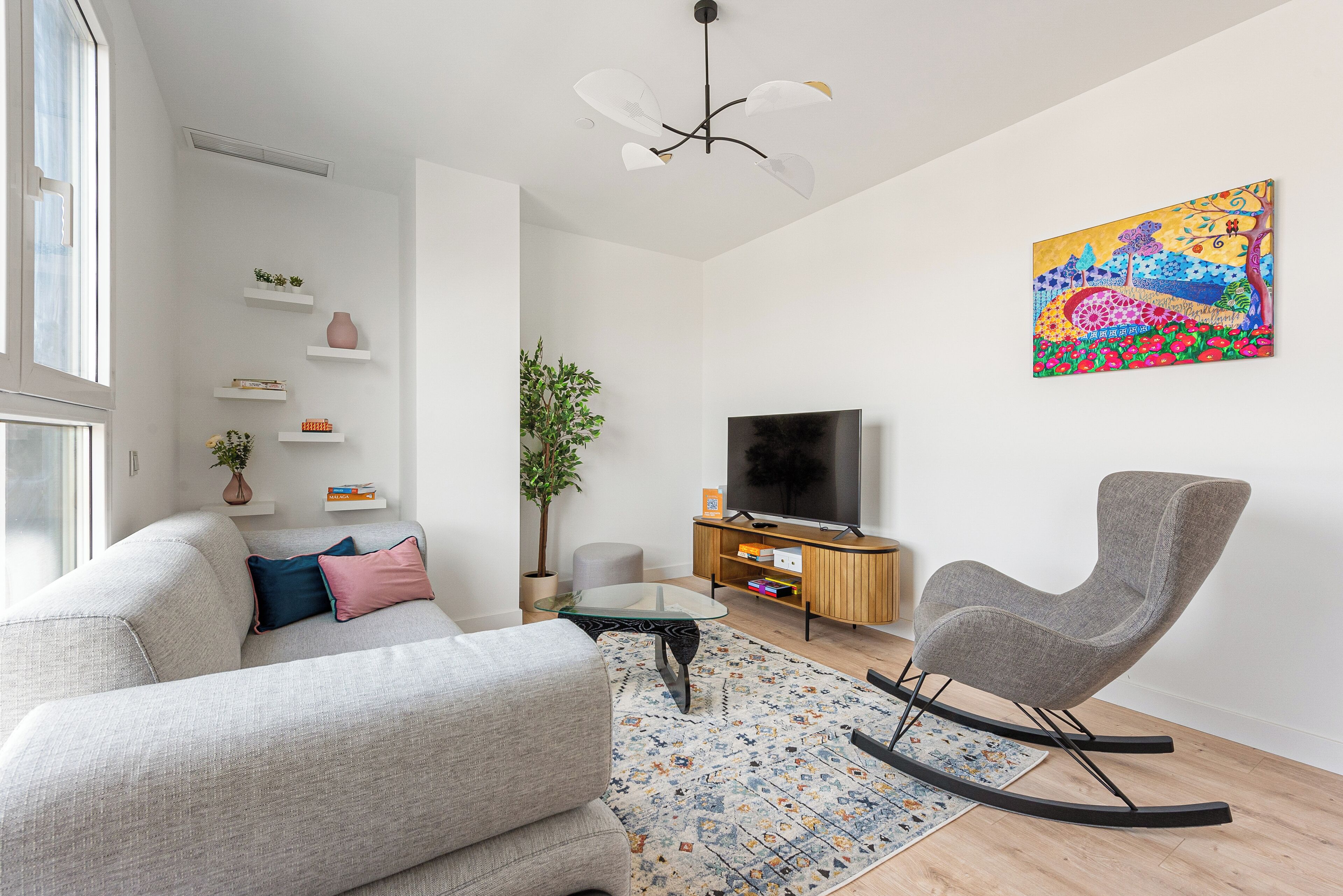 A modern living room with a grey sofa, a glass coffee table, a rocking chair, a flat-screen TV, and a colorful abstract painting on the wall.