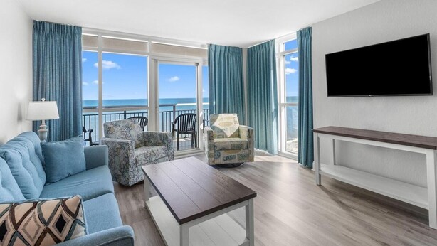 Un soggiorno con divano, poltrona e tavolino, una TV a muro e un balcone con vista sull'oceano.
