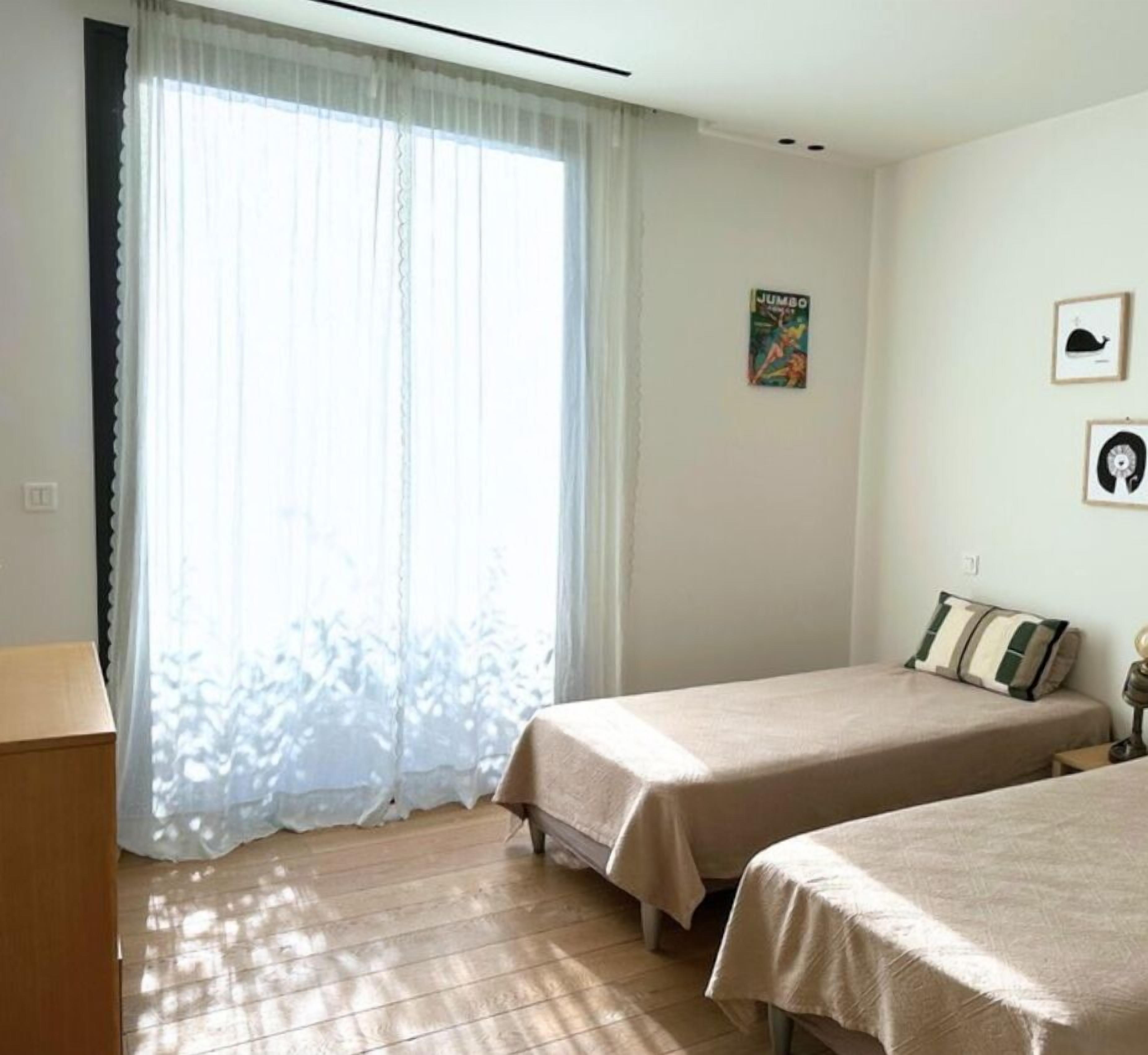 Een kamer met twee bedden, een raam met dunne gordijnen, een poster aan de muur, en een tafeltje met een lamp.