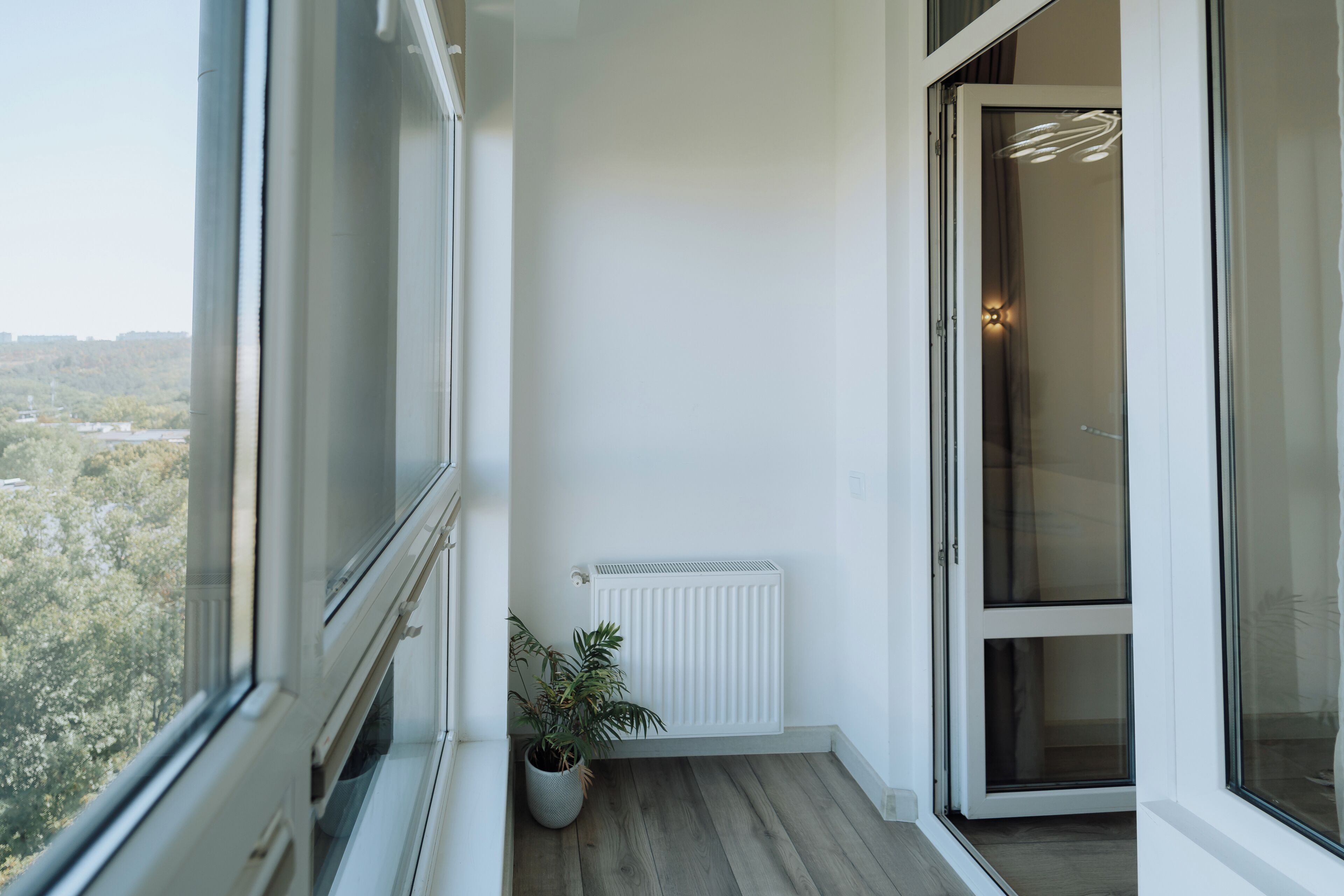 A balcony with a view of trees and buildings, a potted plant, a white radiator, and a sliding glass door.