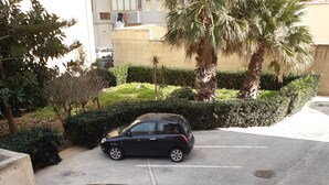 Zona aparcament amb dos cotxes, edifici amb balcons i jardí ben cuidat amb palmeres i arbustos.