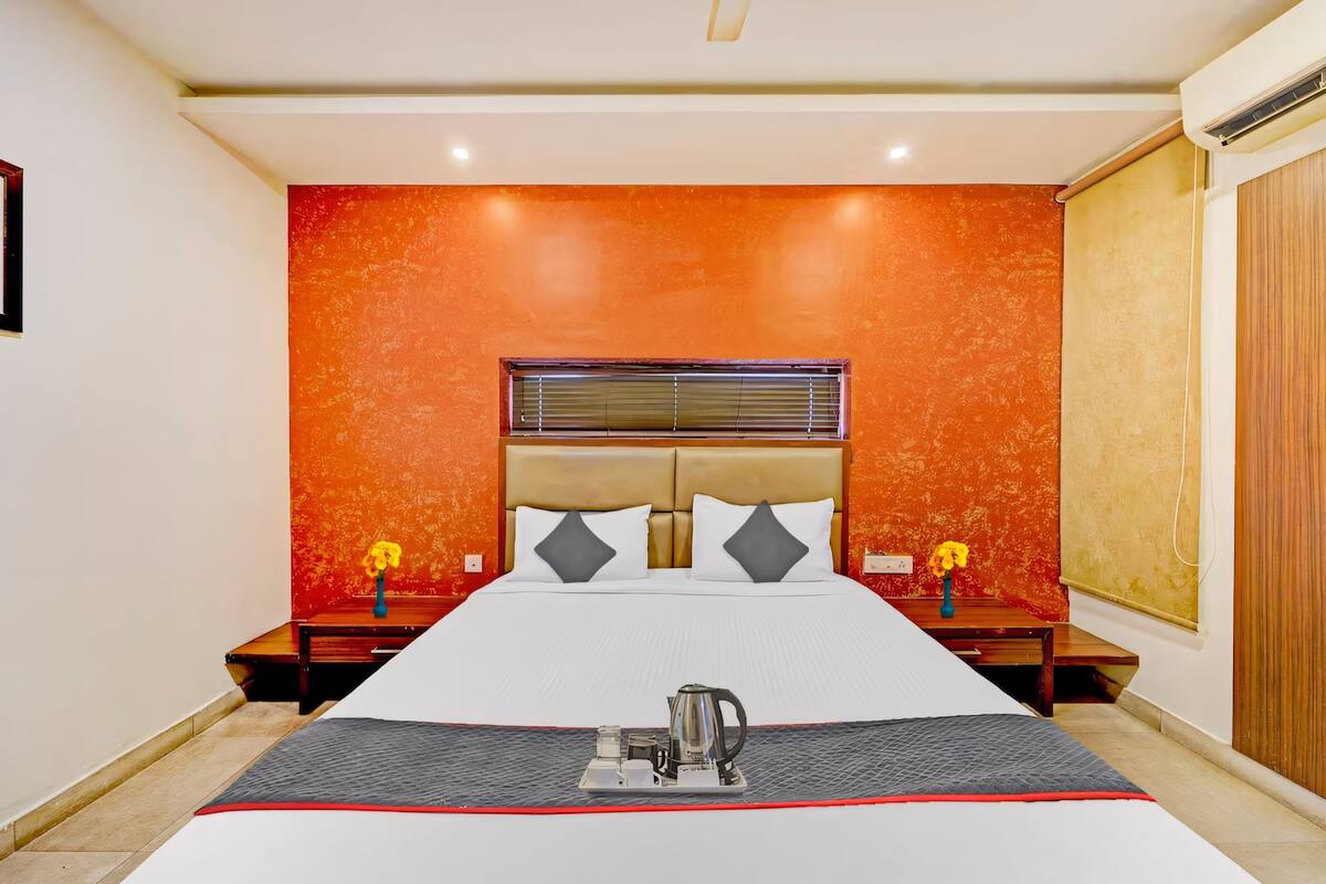 Una habitación de hotel con una cama grande, una mesita de noche con lámpara, una ventana con persianas y una pared con diseño naranja texturizado.