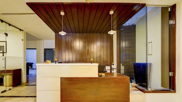 A modern reception area with a wooden ceiling, a white reception desk, and a large mirror. - RG Suites Royapettah (Chennai)
