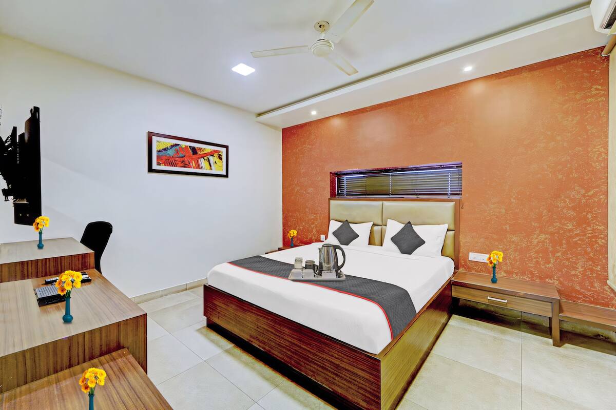 Una habitación de hotel moderna con una cama grande, un escritorio con una silla, una pequeña mesa con flores, un ventilador de techo y una ventana con persianas.