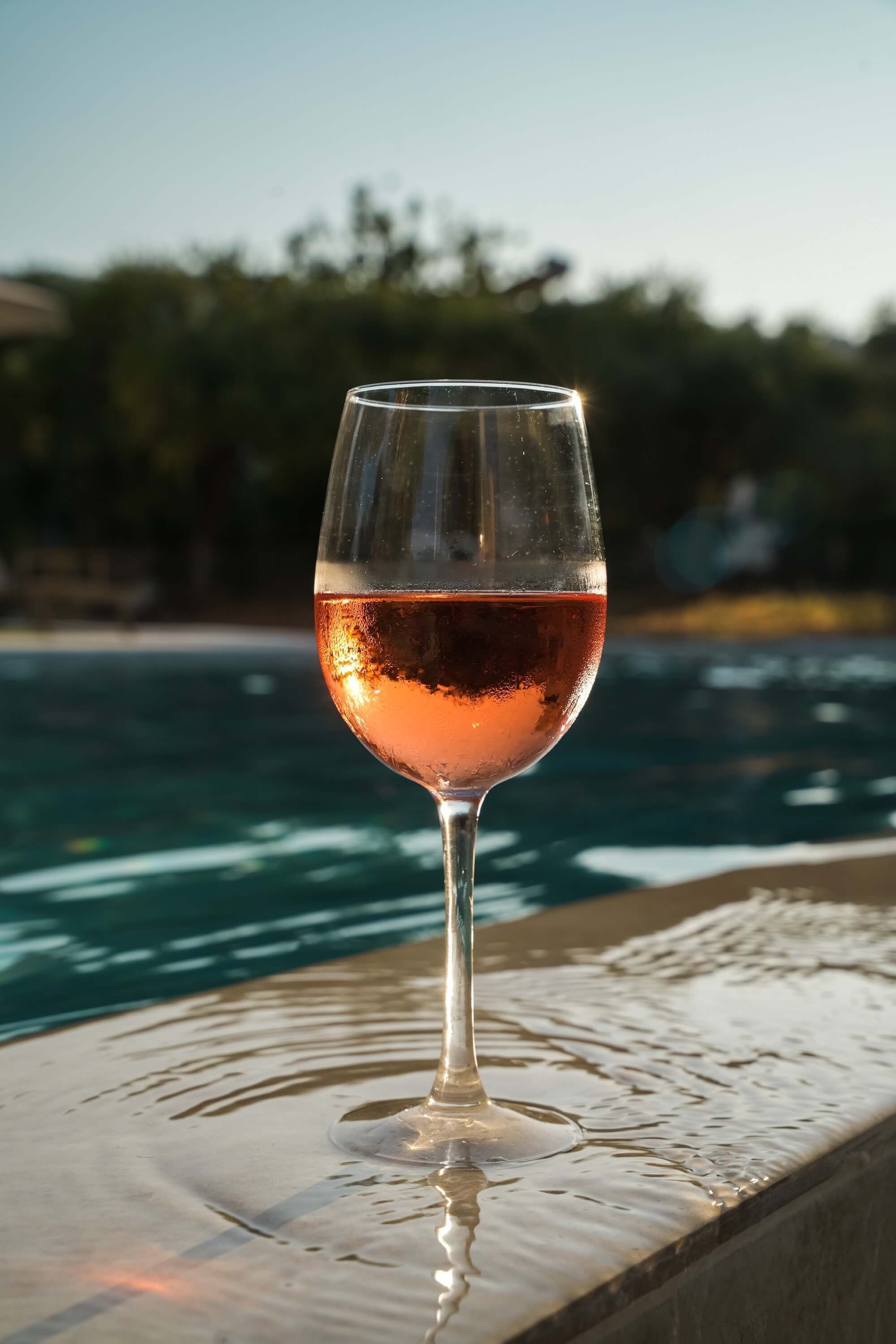 Una copa de vino rosado sobre un alféizar de piedra junto a una piscina.