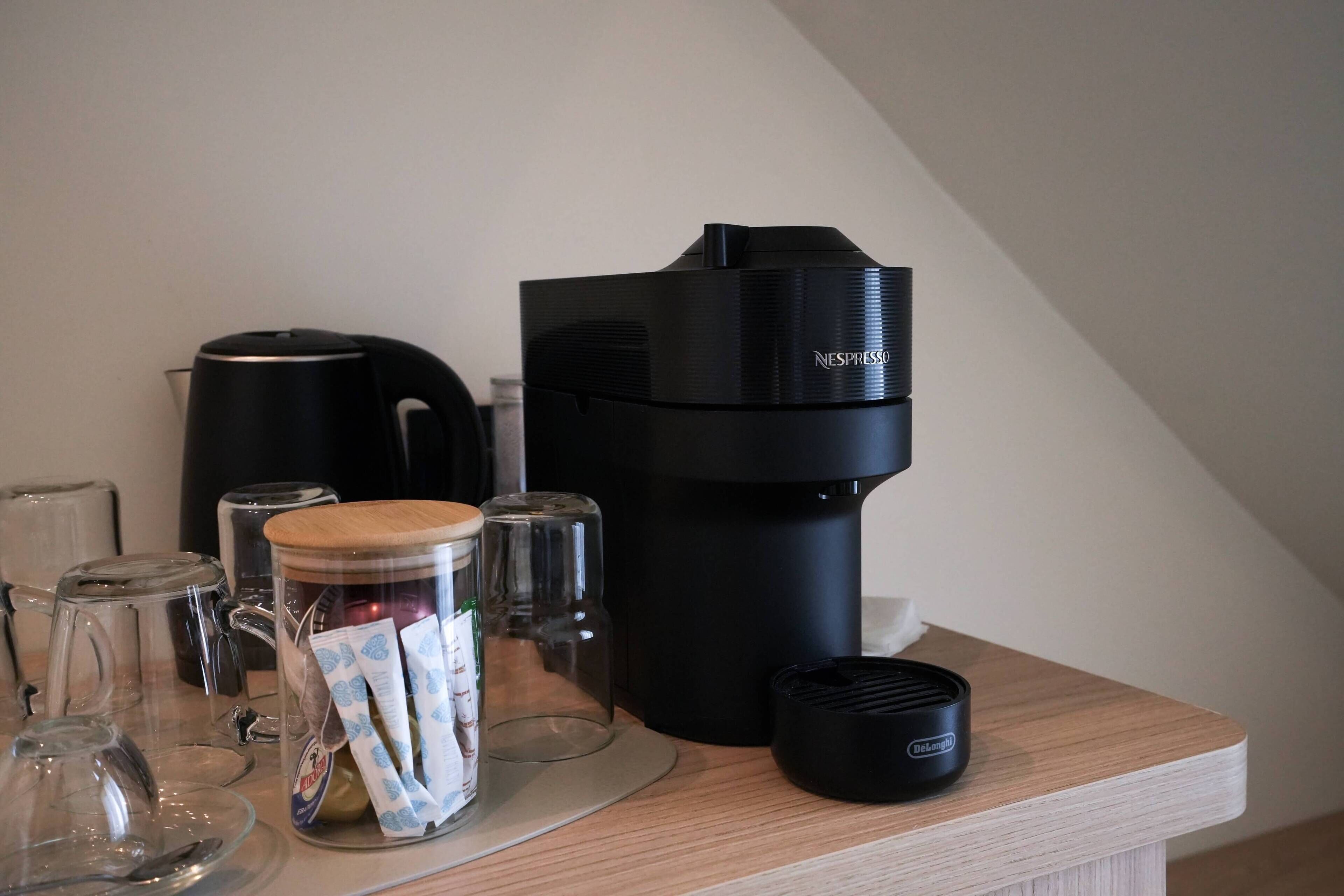 A coffee maker, a glass jar with tea bags, a kettle, and a De'Longhi coffee machine on a wooden countertop.