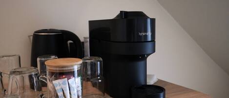 A coffee maker, a glass jar with tea bags, a kettle, and a De'Longhi coffee machine on a wooden countertop.
