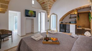 Une pièce spacieuse avec un haut plafond voûté en pierre, un lit agrémenté d’un plateau de fruits et de champagne, une télévision et un coin cuisine.