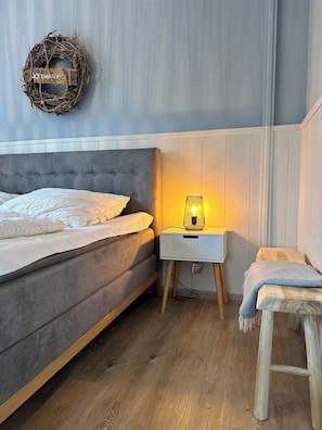 Une chambre à coucher avec un lit recouvert d’un tissu gris, une table de chevet avec une lampe, une petite table en bois et une petite chaise en bois.