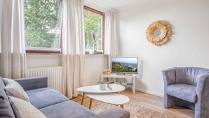Un salon moderne avec un canapé gris, une petite table blanche, un fauteuil gris, un téléviseur et une fenêtre avec des rideaux.