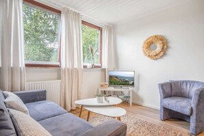 Un salon moderne avec un canapé gris, une petite table blanche, un fauteuil gris, un téléviseur et une fenêtre avec des rideaux.