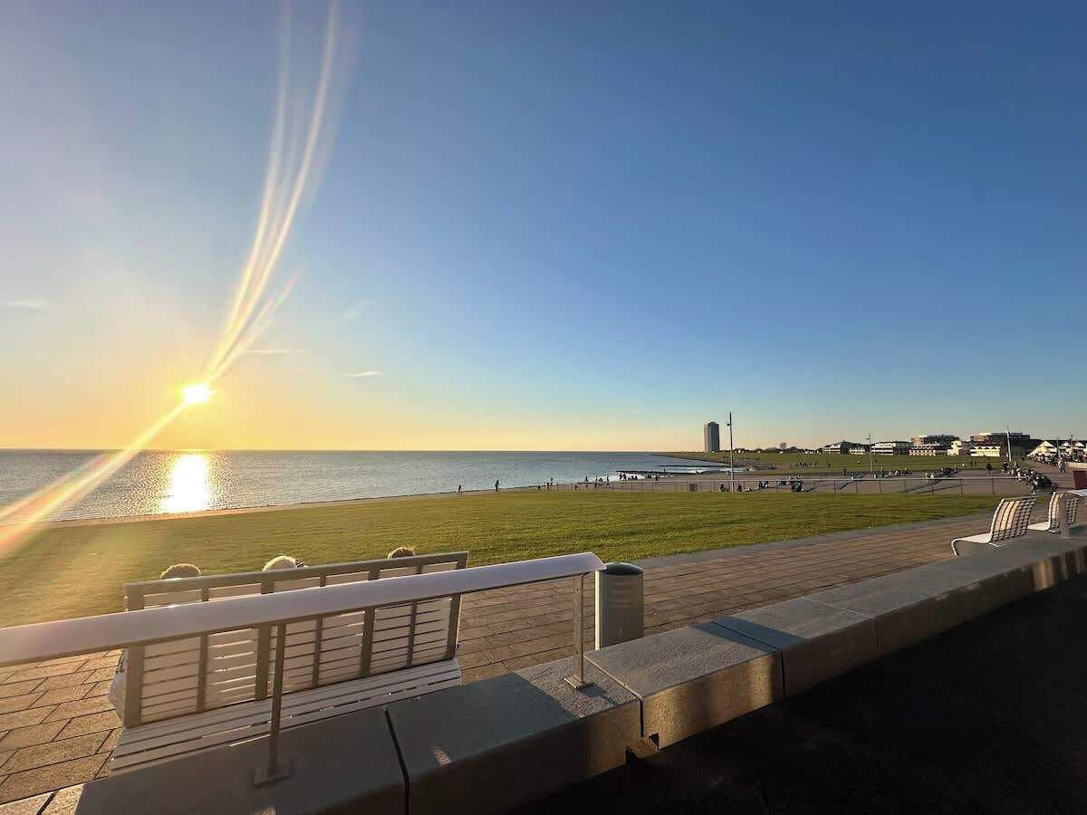 Eine Uferpromenade mit einer Bank, einem Geländer und Blick auf das Meer sowie Gebäude in der Ferne.
