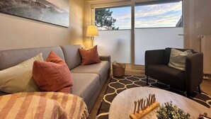 A living room with a sofa, a chair, a coffee table, and a lamp. There is a window with blinds and a view of trees. - Bright apartment at the Family Lagoon w/ balcony (Büsum)