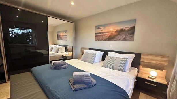 A modern hotel room with a large bed, two bedside tables, a nightstand with a lamp, and a large window with a view of the city. - Bright apartment at the Family Lagoon w/ balcony (Büsum)