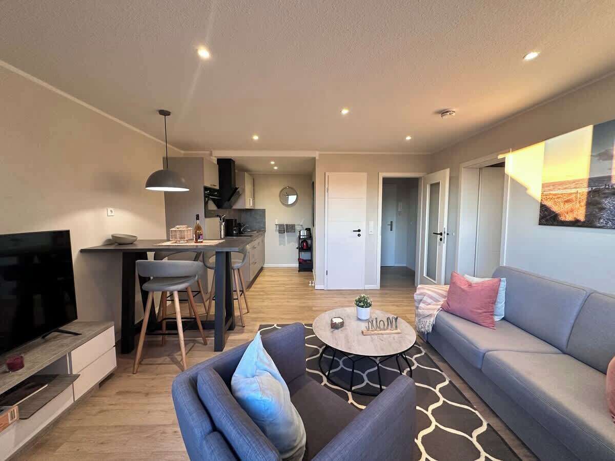 A modern living room with a sofa, coffee table, and TV. There is a kitchen area with a dining table and chairs. The room has a neutral color scheme and a large window.