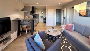 A modern living room with a sofa, coffee table, and TV. There is a kitchen area with a dining table and chairs. The room has a neutral color scheme and a large window. - Bright apartment at the Family Lagoon w/ balcony (Büsum)