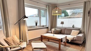 A modern living room with a sofa, armchair, coffee table, and a hanging light fixture. - Ruhige Wohnung am Meer in SPO (Sankt Peter-Ording)