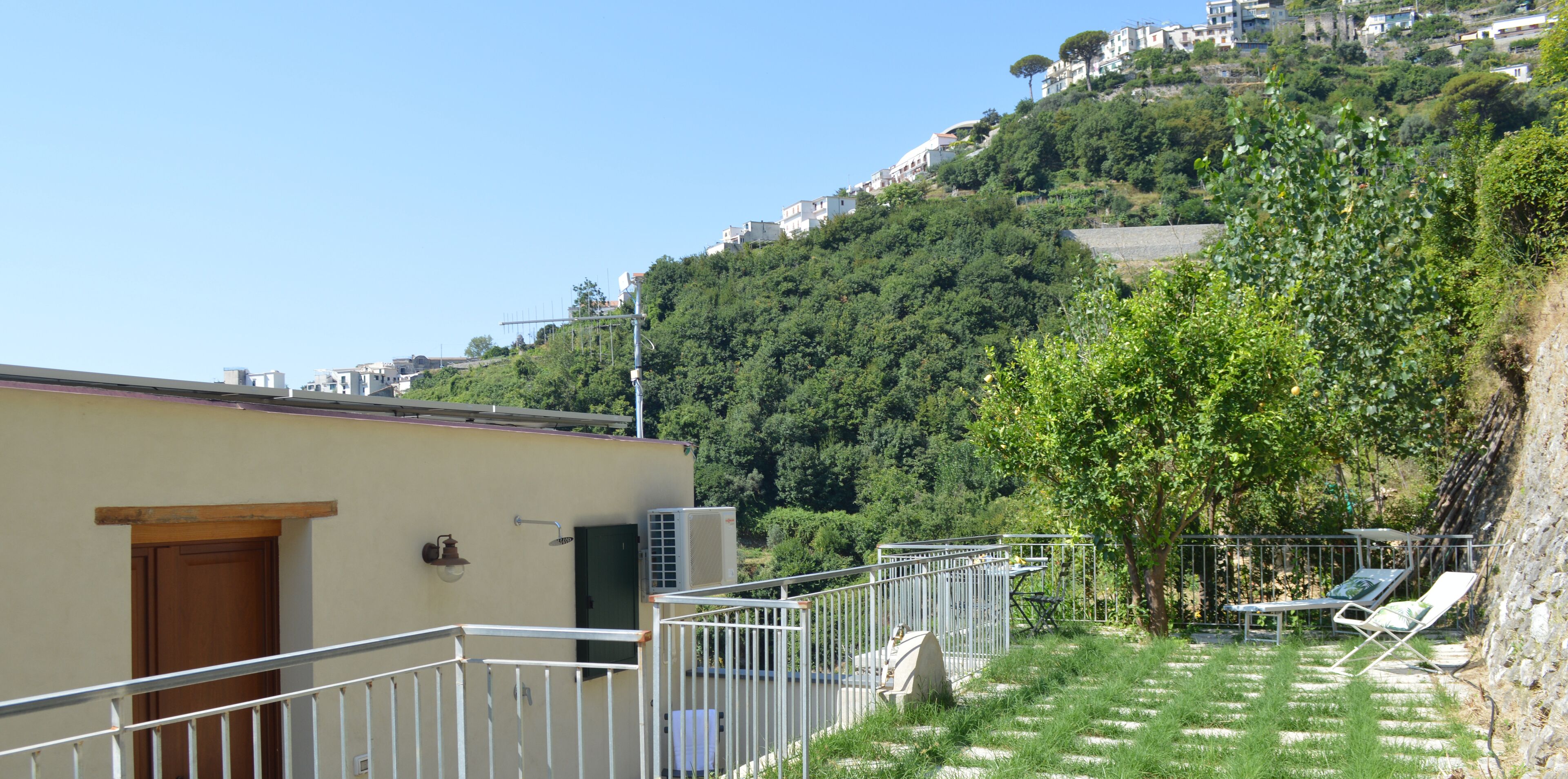 A hillside villa with a balcony, outdoor seating, and a view of the surrounding greenery and buildings.