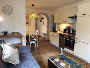 A modern living room with a sofa, a dining table, and a kitchen area with cabinets and appliances. - Strandhütte direkt am Leuchtturm und Strand in SPO (Sankt Peter-Ording)