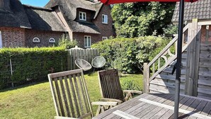 A wooden deck with a table and chairs, a red umbrella, and a brick house with a thatched roof in the background. - Strandhütte direkt am Leuchtturm und Strand in SPO (Sankt Peter-Ording)