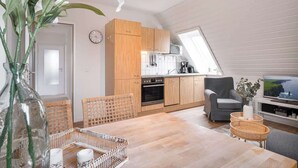 Een moderne keuken met houten kastjes, een eethoek met rieten meubels, en een televisie in de woonkamer.